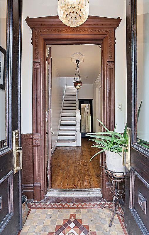 View through the carved Victorian double doors into the entry hall, showing the original tessellated mosaic vestibule floor in geometric patterns, dark walnut wainscoting, a hanging glass chandelier, and the white staircase rising in the background