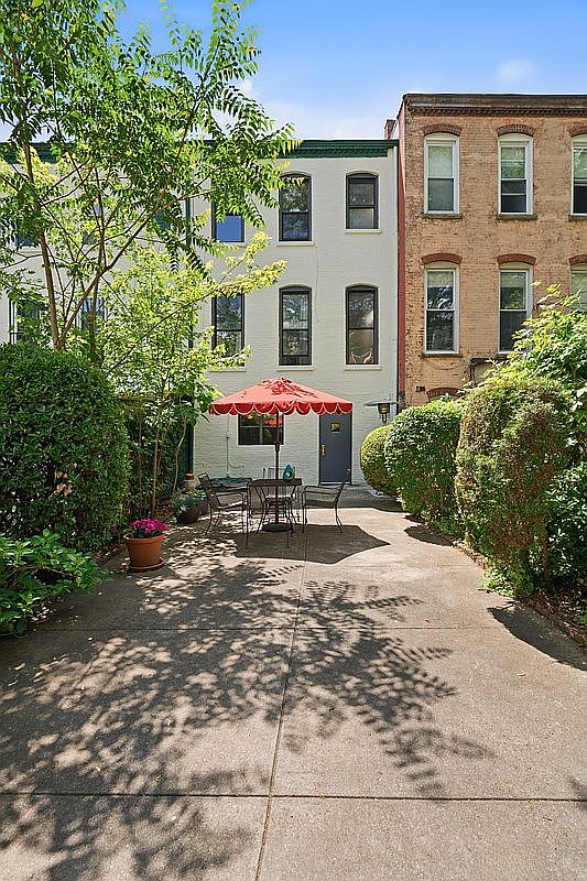 The private back garden at 617 Decatur Street, showing a concrete patio with a red scalloped umbrella and bistro chairs, hedged borders, mature trees overhead, and the brownstone rear facade with painted brick and tall windows