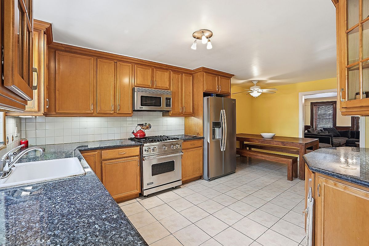 The garden-level chef's kitchen with a stainless Viking range, dark granite counters, warm wood cabinetry, glass-front upper cabinet, tile backsplash, and a benched dining nook in a yellow-walled adjacent room with the living area visible beyond