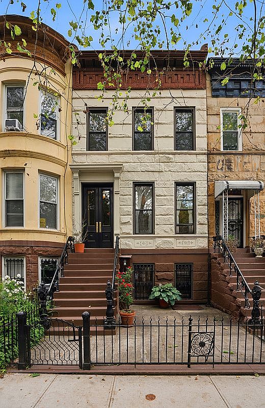 A wider framing of 617 Decatur Street's facade in context, the sandstone front and wrought iron stoop bracketed by the neighboring rowhouses on the tree-lined Bed-Stuy block